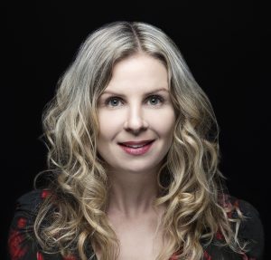 A close-up headshot of a smiling woman with long, wavy blonde hair and light-colored eyes. She is wearing red lipstick and is photographed against a solid black background